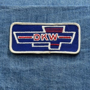 May include: A rectangular patch with a blue background and white border. The patch features the word "DKW" in red letters, with white and red graphic elements. The patch is set against a denim background.