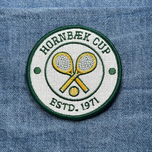 May include: Embroidered patch with the text "HORNBAEK CUP ESTD. 1971" in a circular design. The patch features two crossed tennis rackets and a tennis ball in the center, all in yellow and white, set against a green border. The patch is on a blue denim background.