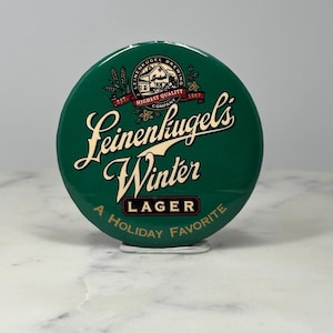 May include: A round, green Leinenkugel's Winter Lager button with white and gold lettering. The button features the brewery's logo and the phrase "A Holiday Favorite."