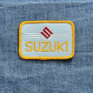 May include: A rectangular patch with the word "SUZUKI" in gold lettering. The patch has a light blue background with a red and white logo and a gold border. The patch is on a blue denim background.