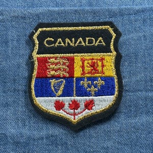 May include: Embroidered patch featuring the word "CANADA" in gold lettering. The patch is shield-shaped with a black border and a design incorporating red, yellow, blue, and white elements, including maple leaves and heraldic symbols. The patch is set against a blue fabric background.