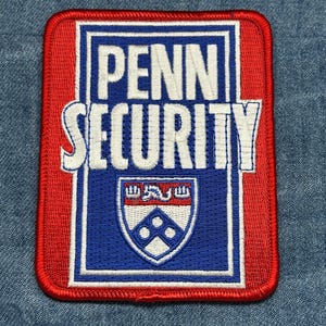 May include: Embroidered patch with the words "PENN SECURITY" in white, set against a red and blue background. The patch features a university crest at the bottom. The patch is rectangular with rounded corners.
