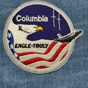 May include: Embroidered patch featuring the word "Columbia" above an eagle and a space shuttle, with the text "ENGLE-TRULY" below. The patch incorporates the American flag's colors and design, with a blue background.