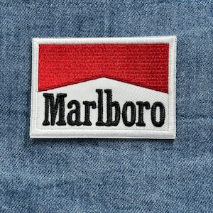 May include: Embroidered patch featuring the Marlboro logo. The patch has a white border, a red top section, a white center, and the word "Marlboro" in black, bold letters. The patch is set against a blue denim background.