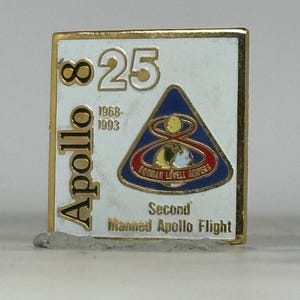 May include: A square, gold-edged pin with a white background. The pin features the word "Apollo" in gold, the numbers "8" and "25", and a blue, red, and gold triangle with text. The text "Second Manned Apollo Flight" is also present.