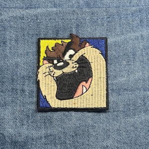 May include: Embroidered patch featuring the Tasmanian Devil cartoon character. The patch has a square shape with a blue and yellow border. The Tasmanian Devil is depicted with a wide open mouth, brown hair, and a tan body.