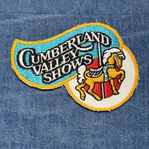 May include: Embroidered patch with the text "CUMBERLAND VALLEY SHOWS" in white, black, and teal. The patch features a carousel horse in yellow, red, and white. The patch is on a blue denim background.