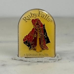 May include: A rectangular pin with a yellow background and gold trim. The pin features the words "Ruby Falls" at the top and "LOOKOUT MT, TN." at the bottom. The center design depicts a figure in red and black robes.