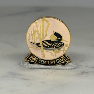 May include: A circular pin with a duck illustration in black and gold, set against a light pink background. The pin features the text "ABA CENTURY CLUB" on a black banner at the bottom.