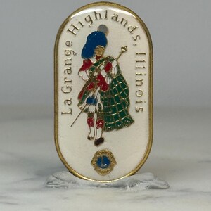 May include: An oval-shaped pin with a gold border and a white background. The pin features a Scottish Highlander in traditional attire, with the text "La Grange Highlands, Illinois". A blue and gold emblem is at the bottom.
