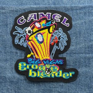 May include: Embroidered patch with the text "CAMEL" in purple, and "BIZ VEGAS Groove blender" in blue and yellow. The patch features a cartoon image of a yellow object with palm trees and musical notes on a black background.