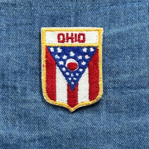 May include: Embroidered patch featuring the Ohio state flag design. The patch has a shield shape with a white border and the word "OHIO" in red. The flag design includes red and white stripes, a blue triangle with white stars, and a red circle.
