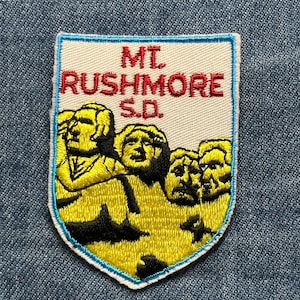 May include: Embroidered patch featuring Mount Rushmore in yellow and black, with the text "MT. RUSHMORE S.D." in red. The patch has a white background with a blue border, and is set against a denim background.