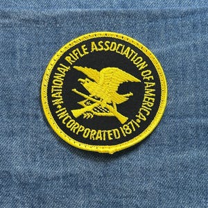 May include: A round patch with a black center and yellow border. The text "NATIONAL RIFLE ASSOCIATION OF AMERICA INCORPORATED 1871" is in yellow around the edge. A yellow eagle and crossed rifles are in the center.