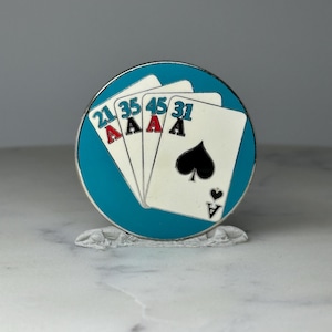 May include: A round, teal-colored poker chip featuring four playing cards. The cards are white with black and red suits, and the numbers 21, 35, 45, and 31 are printed on them. The chip is resting on a white, textured surface.