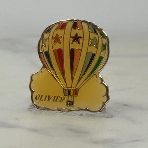 May include: A gold-colored hot air balloon pin with a colorful design. The balloon features red, orange, yellow, green, and blue stripes, with star accents. The word "OLIVIER" is written in gold on the cloud.