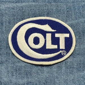 May include: Oval-shaped patch with the word "COLT" in white, embroidered on a blue background. The patch has a white border and is set against a denim fabric backdrop. The registered trademark symbol is visible.