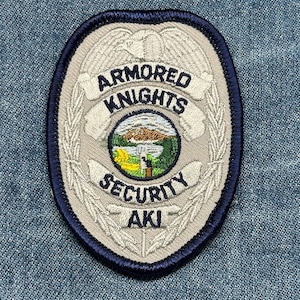 May include: Embroidered patch with a shield shape, featuring the text "ARMORED KNIGHTS SECURITY AKI." The patch has a gray background with a blue border and a central image of a landscape. The design includes an eagle and laurel leaves.