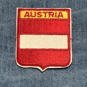 May include: Embroidered patch featuring the Austrian flag design. The patch is shield-shaped with a red background, a white horizontal stripe, and a yellow banner at the top with the word "AUSTRIA". The patch is on a blue denim background.