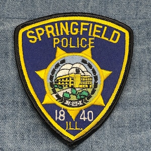 May include: A Springfield Police patch with a blue background and yellow trim. The patch features the words "SPRINGFIELD POLICE" in yellow, a central emblem with a building, and the numbers "18 40" with "ILL" below.