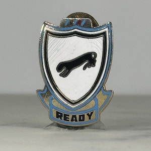 May include: A vintage metal pin with a shield design. The pin features a white background with a black jaguar leaping across it. The shield is outlined in black and blue, with the word "READY" in gold lettering.