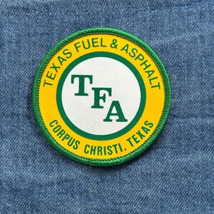 May include: Circular patch with a yellow background and green border. The text "TEXAS FUEL & ASPHALT" arches around the top, and "CORPUS CHRISTI. TEXAS" arches around the bottom. The center features "TFA" in green on a white circle.