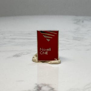 May include: A rectangular red pin with the text "Novell CNE" in white. The pin has a gold border and a stylized design at the top. The pin is resting on a beige, clay-like material.