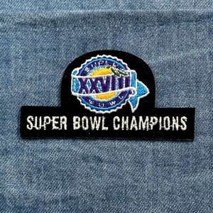 May include: A black patch with white embroidered text reading "SUPER BOWL CHAMPIONS." Above the text is a circular patch with the text "SUPER BOWL" and the Roman numeral "XXVIII" in blue and yellow.