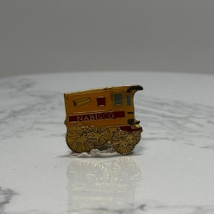 May include: A gold and red Nabisco pin in the shape of a stagecoach. The pin has a yellow body with red accents and the word "NABISCO" in red lettering. The wheels are gold.