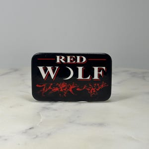 May include: A rectangular black pin with the words "RED WOLF" in white and red lettering. Below the text is a red graphic of wolves. The pin has a glossy finish.