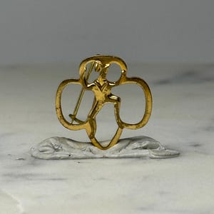 May include: A gold-colored pin featuring a stylized figure within a trefoil shape. The pin has a smooth, polished finish and is set against a neutral background. The design is a classic emblem, likely representing an organization or group.