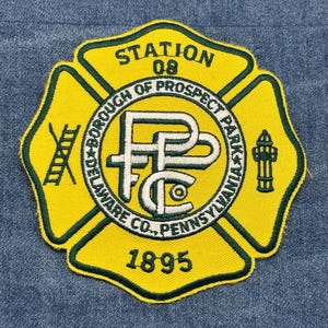 May include: A yellow and green embroidered patch featuring the text "STATION 08" and "BOROUGH OF PROSPECT PARK, DELAWARE CO., PENNSYLVANIA 1895". The patch has a Maltese cross design with a ladder, a fire hydrant, and the letters "FPC".