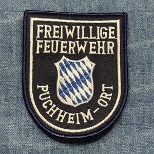 May include: A navy blue and white embroidered patch with the text "FREIWILLIGE FEUERWEHR PUCHHEIM-ORT". The patch features a shield with a blue and white diamond pattern.