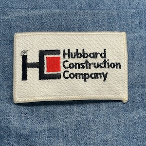 May include: A rectangular white patch with the text "Hubbard Construction Company" in black. The logo features a black "H" with a red square in the middle. The patch is on a blue denim background.