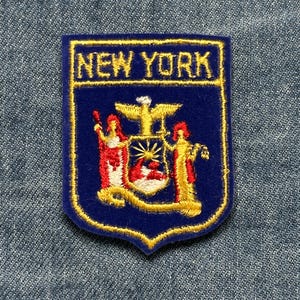 May include: A blue embroidered patch with the words "NEW YORK" in gold lettering. The patch features the New York state coat of arms, including figures, an eagle, and a shield, all outlined in gold.