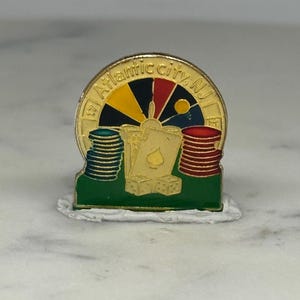 May include: A gold-colored pin featuring a casino theme. The pin includes stacks of poker chips, playing cards, and dice on a green base. The top of the pin has a circular design with the text "Atlantic City, NJ".