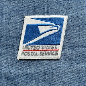 May include: A rectangular patch featuring the United States Postal Service logo, a blue and white stylized eagle. The text "UNITED STATES POSTAL SERVICE" is embroidered below the logo in gray and red. The patch is set against a blue denim background.