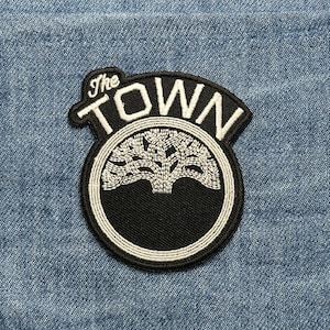 May include: Black and white embroidered patch with the text "The TOWN" in a stylized font. The patch features a tree design within a circular emblem, set against a denim background. The patch is likely for apparel.