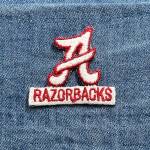 May include: Embroidered patch featuring a white and red letter "A" above a rectangular patch with the word "RAZORBACKS" in red. The patches are on a blue denim background.