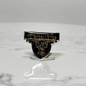 May include: A silver and black Raiders pin with the words "COMMITMENT TO EXCELLENCE" above the team logo. The logo is a shield with a silver outline and a black interior, featuring a silver Raider head.