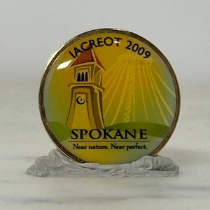 May include: A round, gold-toned pin with a colorful design. The pin features a clock tower, the text "IACREOT 2009" and "SPOKANE" with the tagline "Near nature. Near perfect." The background is yellow and green.