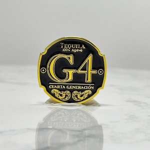 May include: A black and gold metal pin with the text "TEQUILA 100% Agave" at the top. The pin features a large "G4" in yellow, with "CUARTA GENERACIÓN" below. Ornate gold detailing is present.