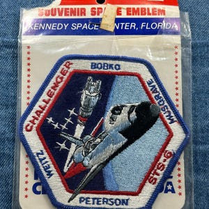 May include: A souvenir patch featuring a space shuttle and the text "Challenger" and "STS-6". The patch is in a hexagonal shape with a blue and red color scheme, and the text "Kennedy Space Center, Florida" is at the top.