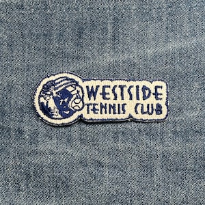 May include: A rectangular patch with the text "WESTSIDE TENNIS CLUB" in white, outlined in blue. The left side features a blue tennis ball and a stylized panther head. The patch is on a blue denim background.