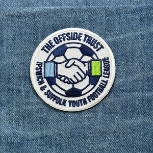 May include: A circular patch with the text "THE OFFSIDE TRUST" and "IPSWICH & SUFFOLK YOUTH FOOTBALL LEAGUE" in a navy blue font. The patch features a soccer ball design with two hands shaking in the center. The background is denim.