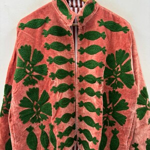 May include: Coral-colored jacket with green embroidered floral and abstract designs. The jacket has a collarless design and a striped lining. The embroidery is detailed and covers the front and sleeves of the jacket. The jacket is hanging on a wooden hanger.