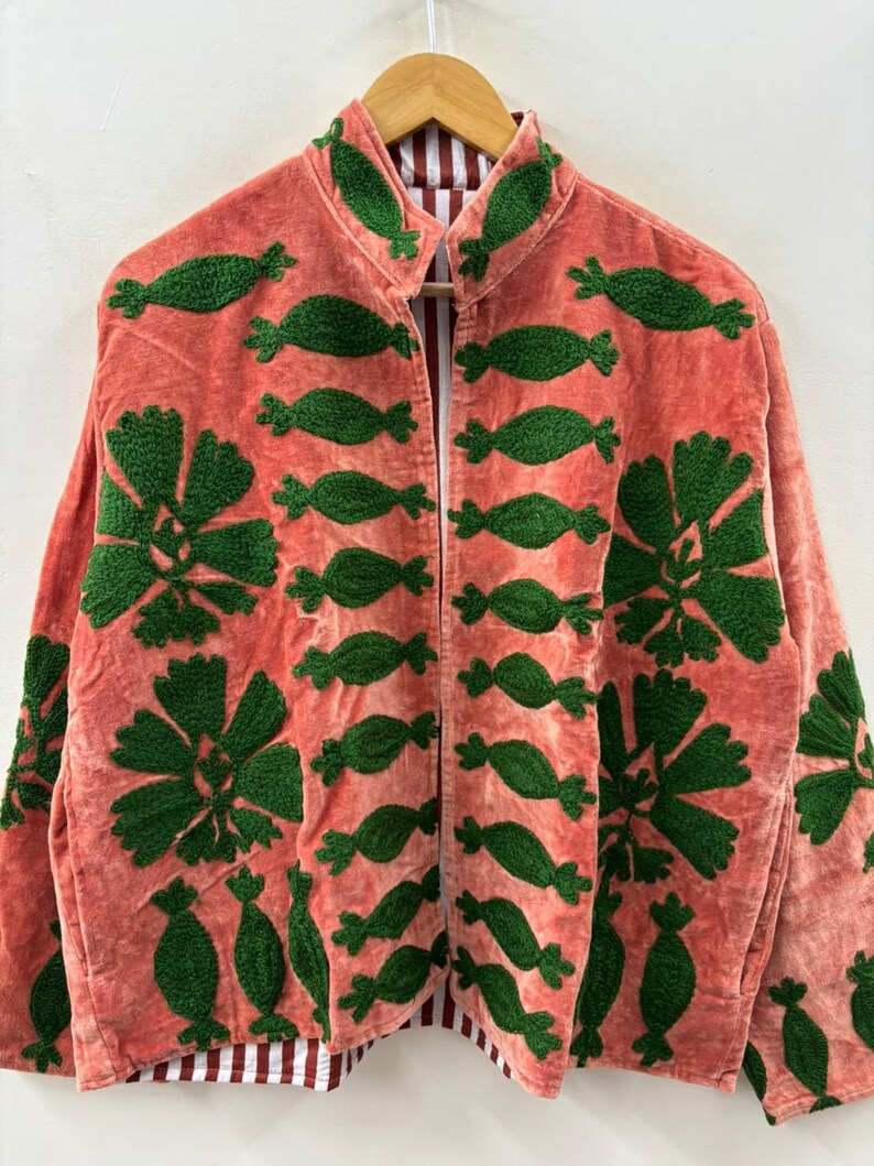 May include: Coral-colored jacket with green embroidered floral and abstract designs. The jacket has a collarless design and a striped lining. The embroidery is detailed and covers the front and sleeves of the jacket. The jacket is hanging on a wooden hanger.