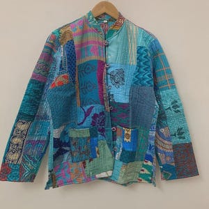 Könnte beinhalten: Eine Patchwork-Jacke in Blau-, Türkis- und Grüntönen mit Stehkragen und Knopfverschluss. Die Jacke besteht aus verschiedenen Stoffmustern und -strukturen, wodurch ein einzigartiges, gestepptes Design entsteht. Sie hängt an einem Holzbügel.