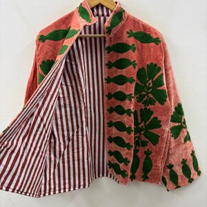 May include: Coral-colored jacket with green embroidered floral and leaf designs. The jacket features a mandarin collar and is lined with red and white striped fabric. The jacket is hanging on a wooden hanger.