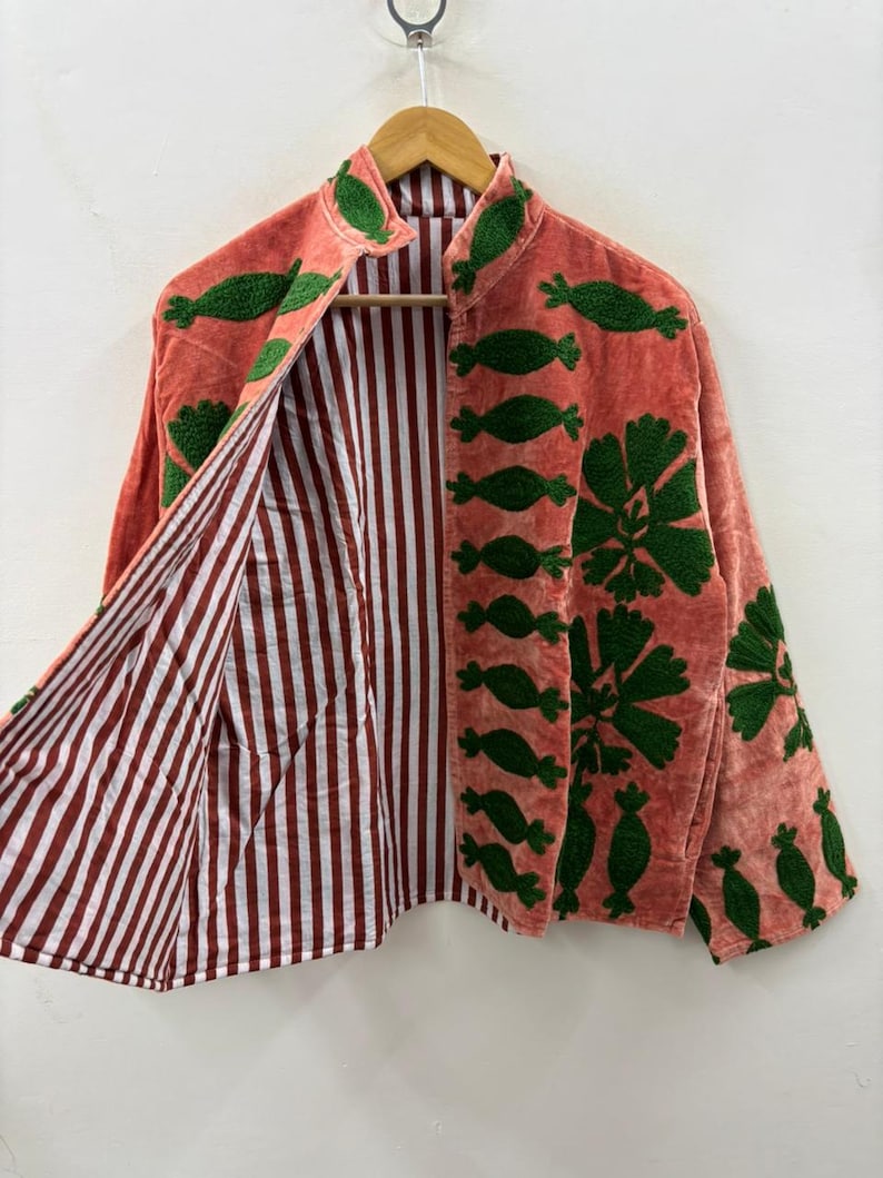 May include: Coral-colored jacket with green embroidered floral and leaf designs. The jacket features a mandarin collar and is lined with red and white striped fabric. The jacket is hanging on a wooden hanger.